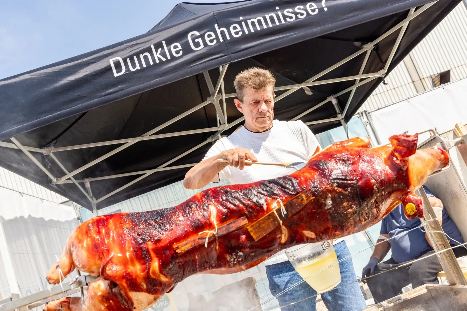 Ein Griller bestreicht das Spanferkel am Spieß unter dem Pavillon