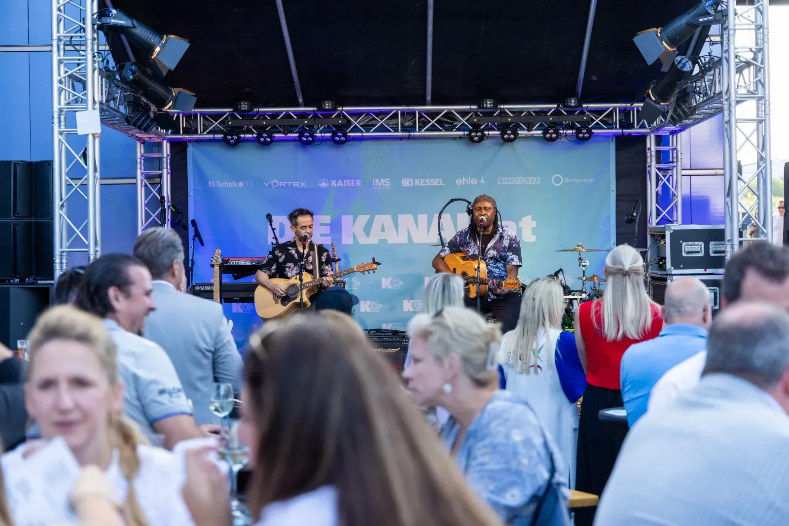 Zwei Musiker auf der Bühne, im Vordergrund die Gäste an den Tischen