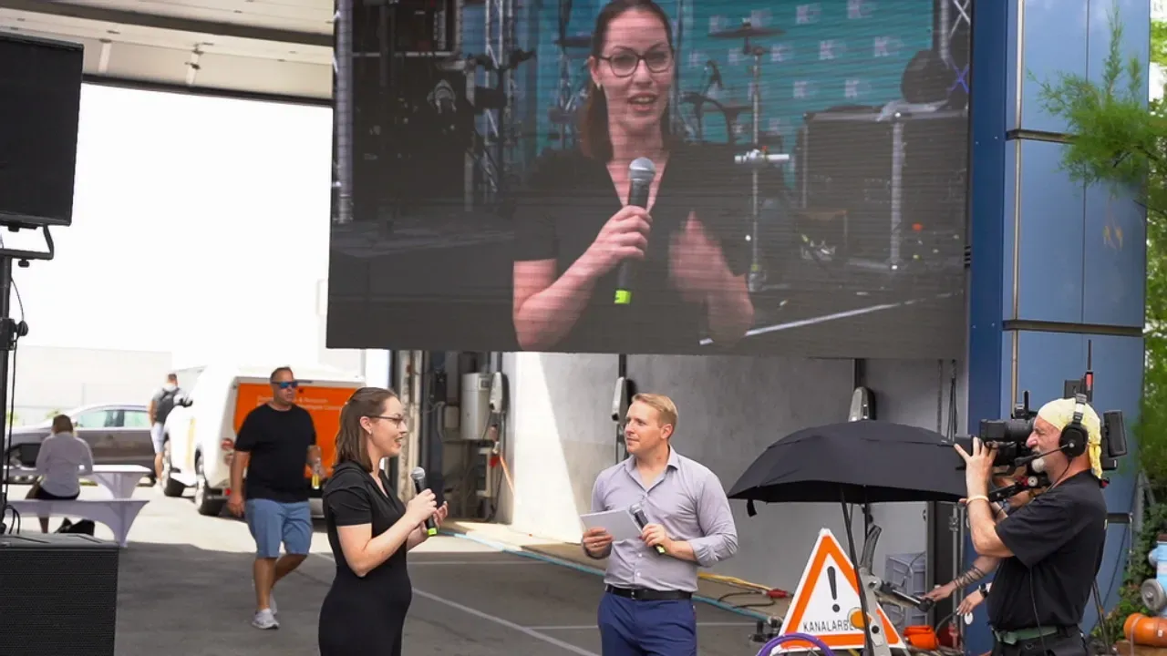Moderationsduo im Freien, dahinter die eigene Übertragung auf der LED-Wand